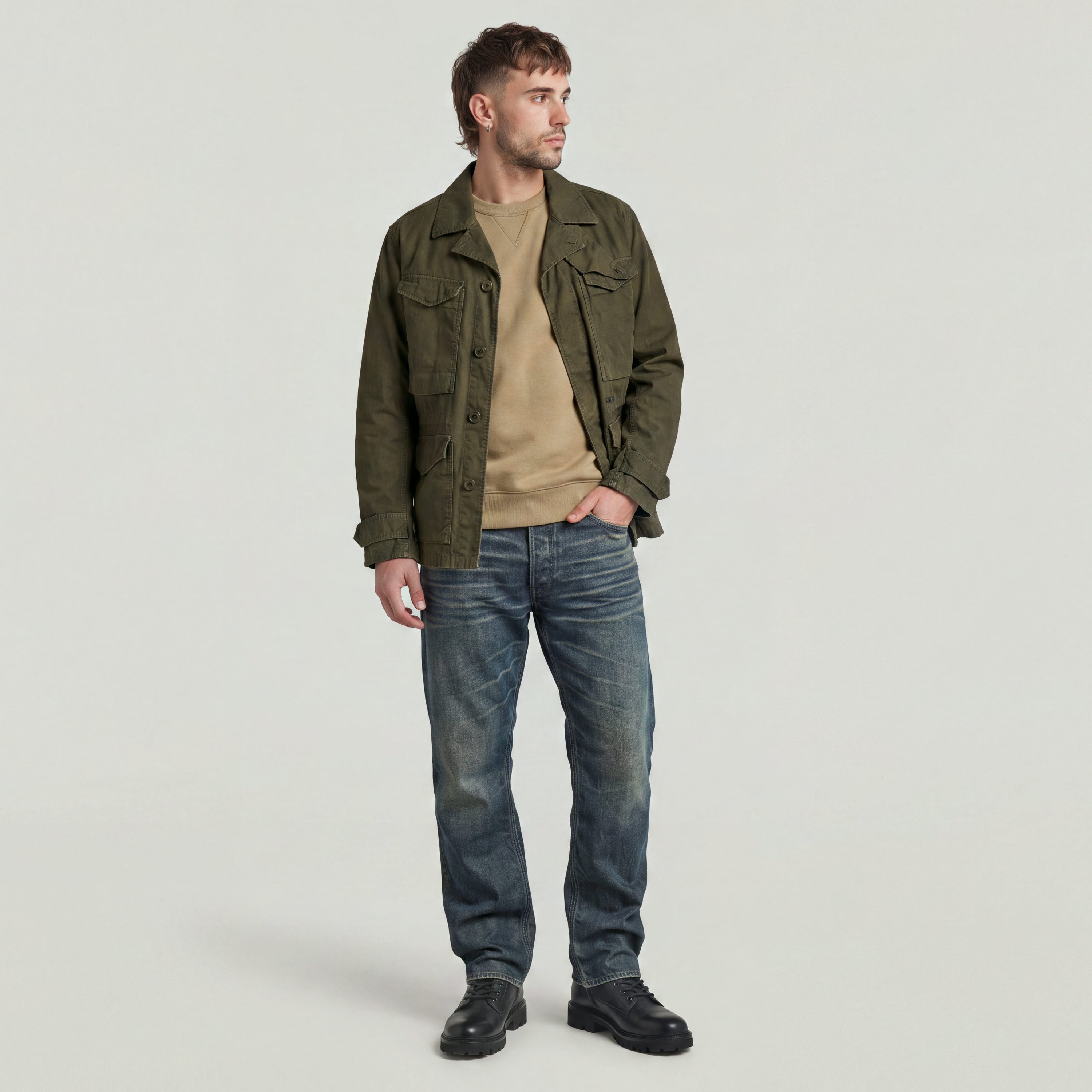 Hombre usando un Premium Core Sweater bajo una chaqueta verde, con jeans y botas, posando en un fondo neutral.