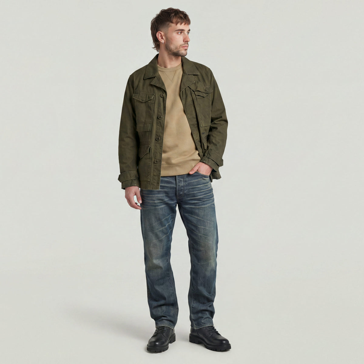 Hombre usando un Premium Core Sweater bajo una chaqueta verde, con jeans y botas, posando en un fondo neutral.