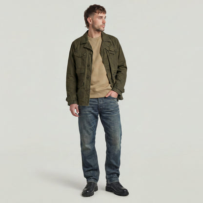 Hombre usando un Premium Core Sweater bajo una chaqueta verde, con jeans y botas, posando en un fondo neutral.