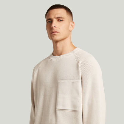 Hombre luciendo un jersey de punto con bolsillo acanalado, estilo raglan rib pocket knit, corte regular y mangas largas.