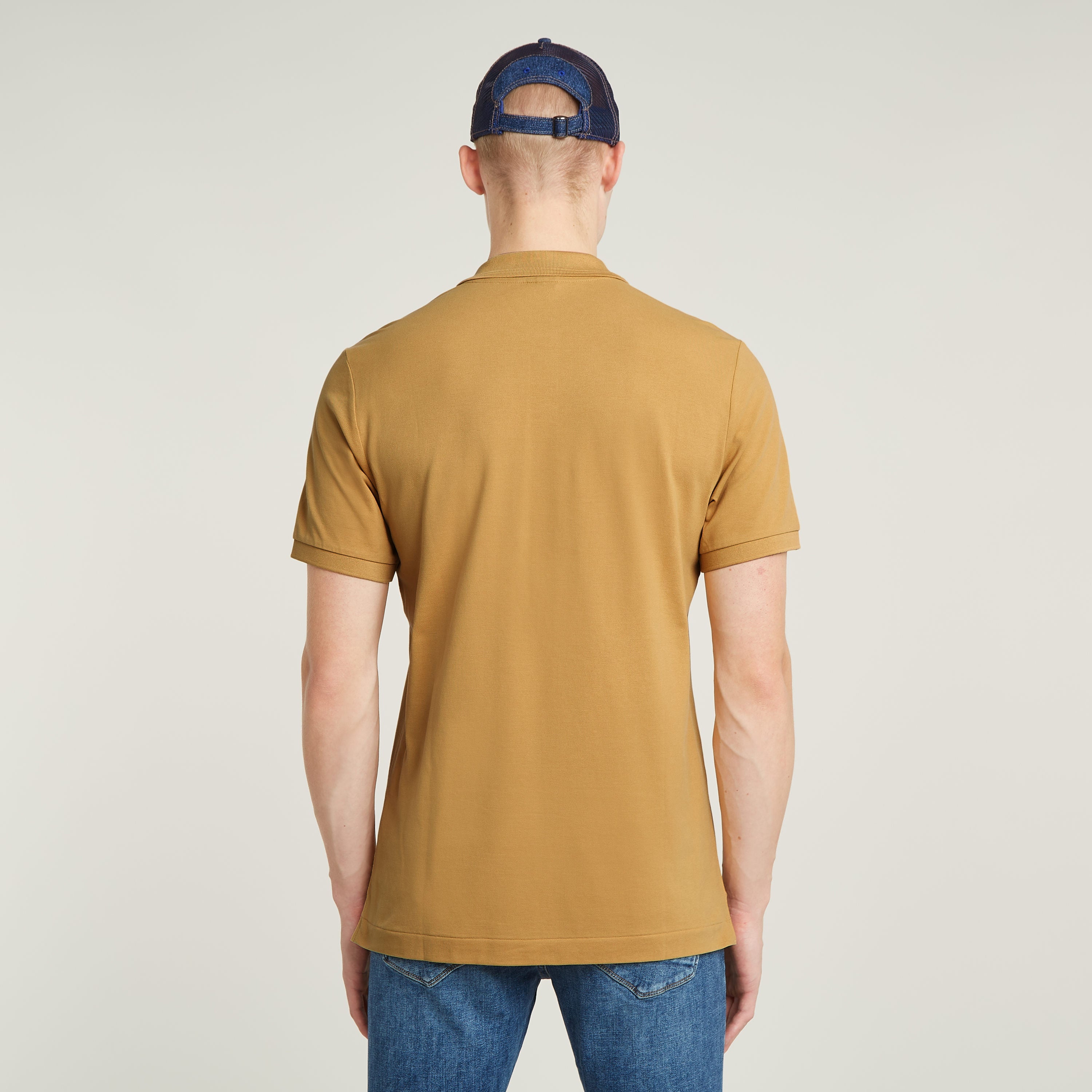 Modelo vistiendo el polo Dunda Slim en color marrón, vista trasera con gorra azul.