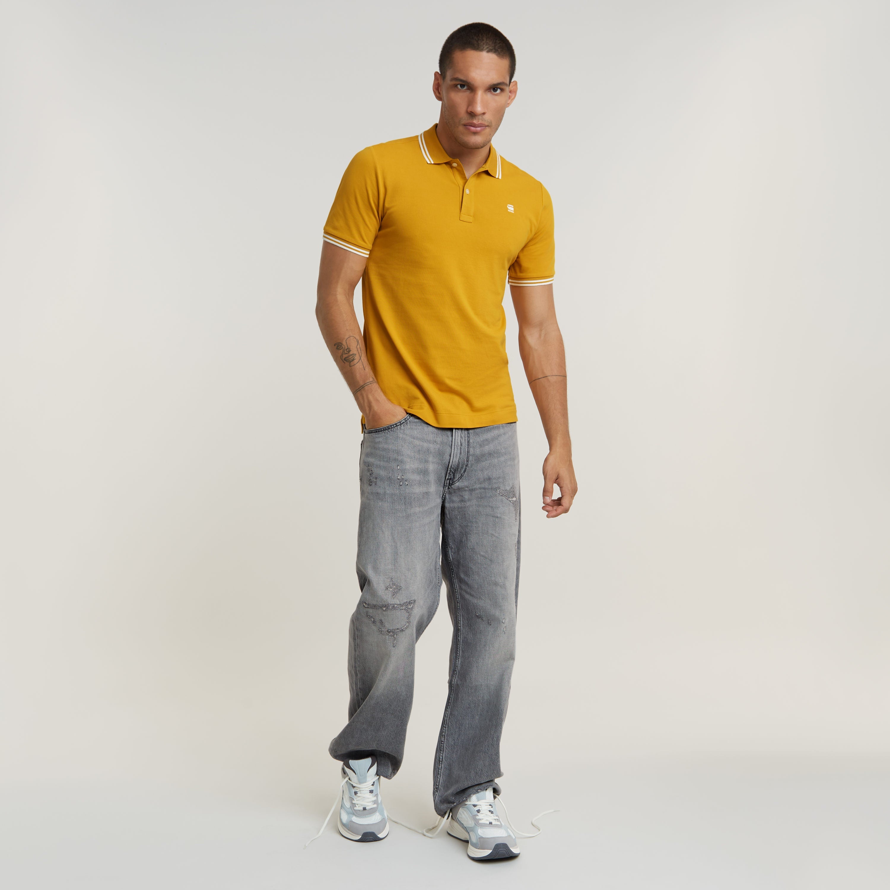 Hombre usando una camiseta polo Dunda Slim Stripe amarilla con detalles de rayas, combinada con jeans y zapatillas.