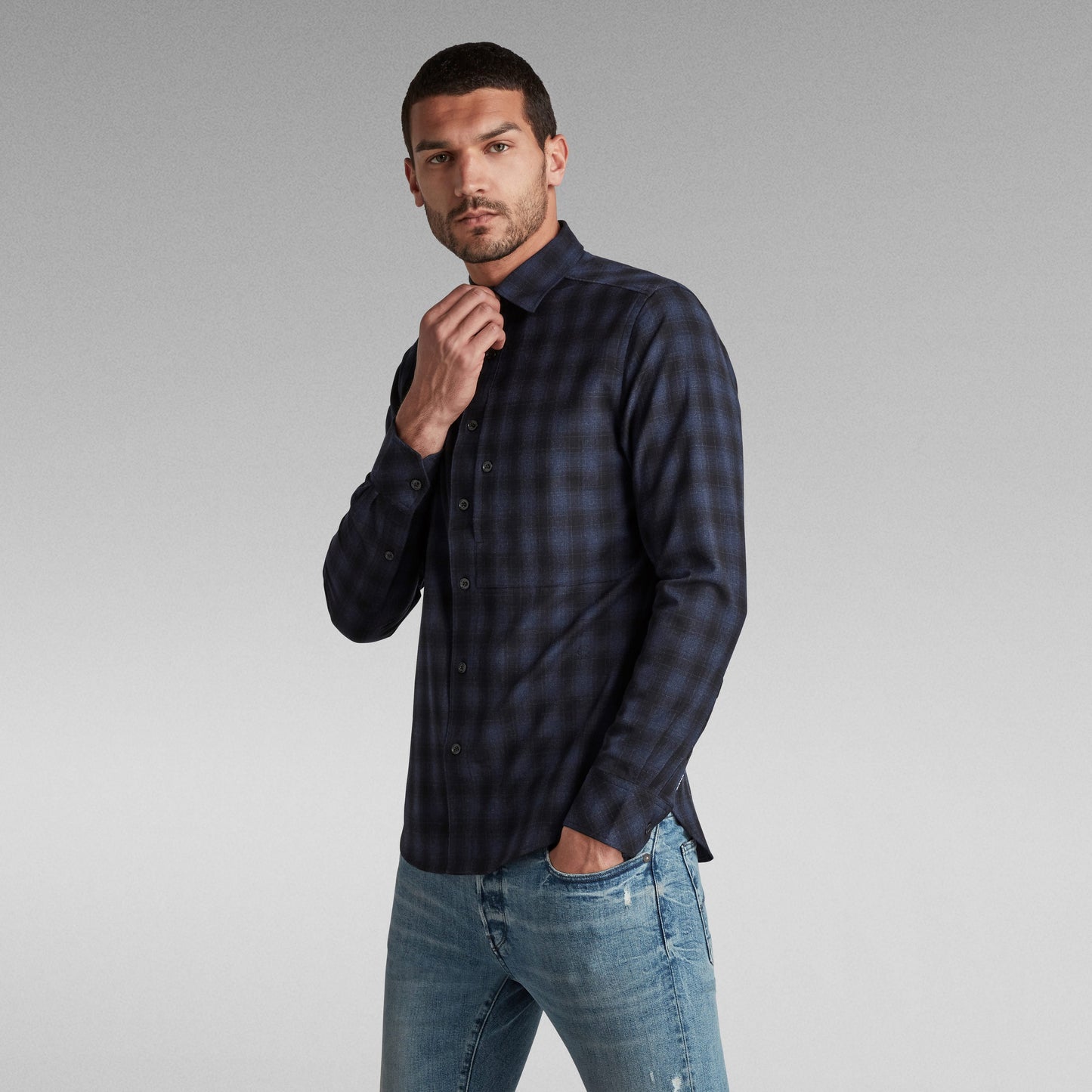 Hombre luciendo la camisa slim fit G Star en patrón de cuadros, combinada con jeans y fondo neutro.
