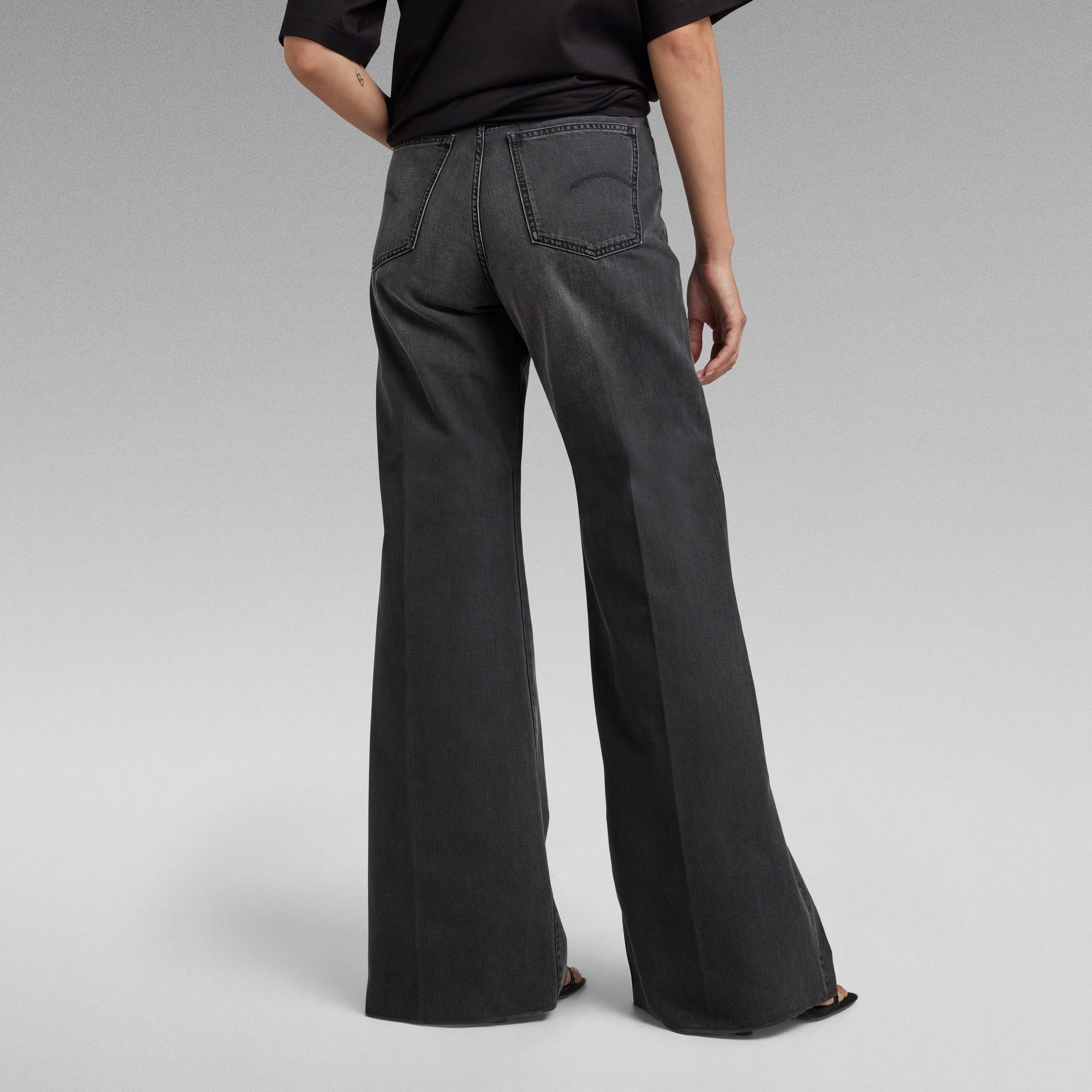 Mujer usando jeans Deck ultra high wide leg jeans con pernera ancha desde atrás en fondo gris.