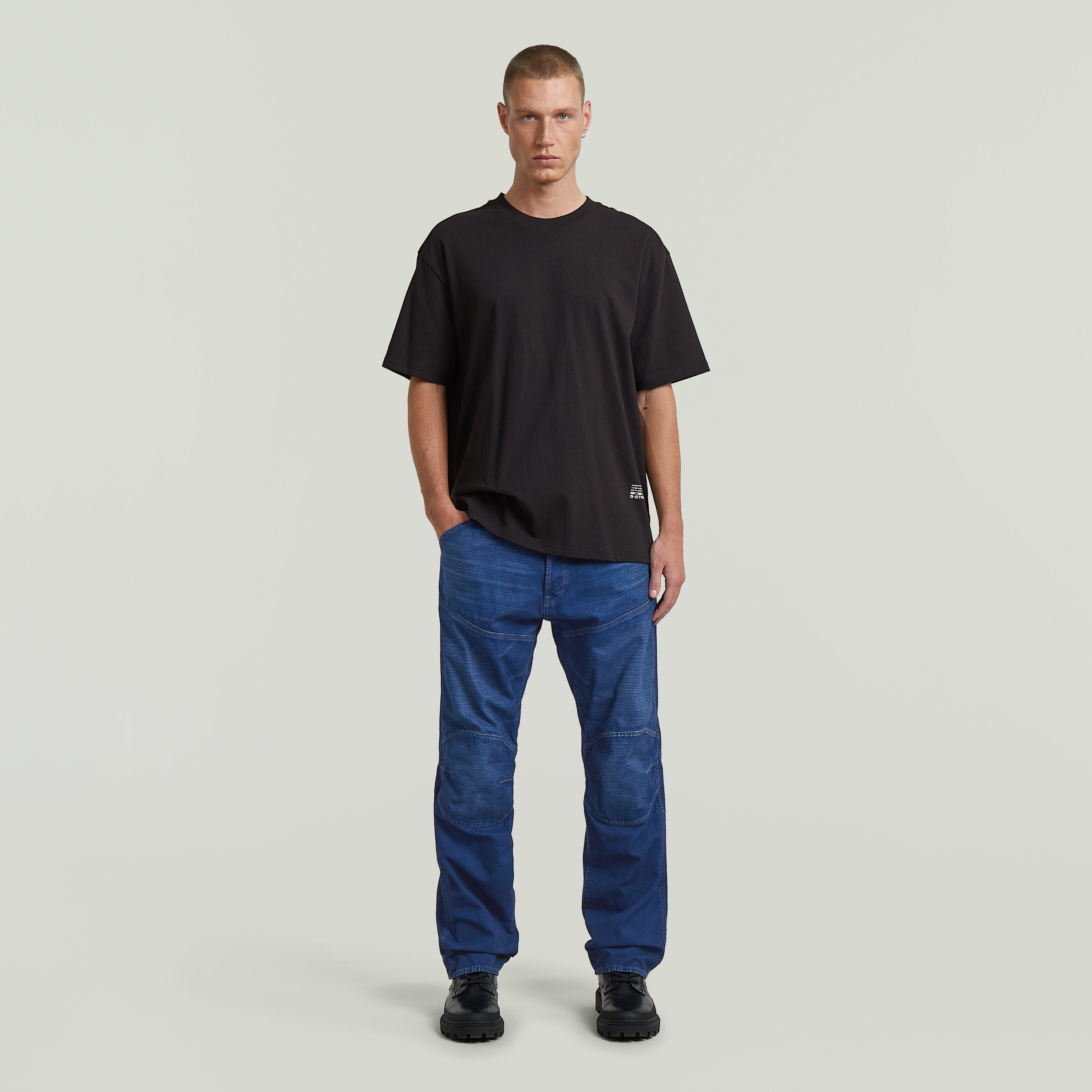 Hombre vistiendo jeans G-STAR Elwood regular, camiseta negra y zapatos negros, fondo claro.