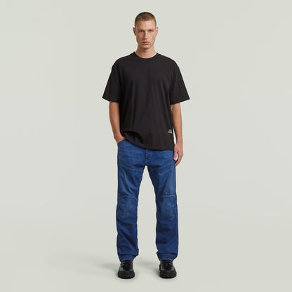 Hombre vistiendo jeans G-STAR Elwood regular, camiseta negra y zapatos negros, fondo claro.