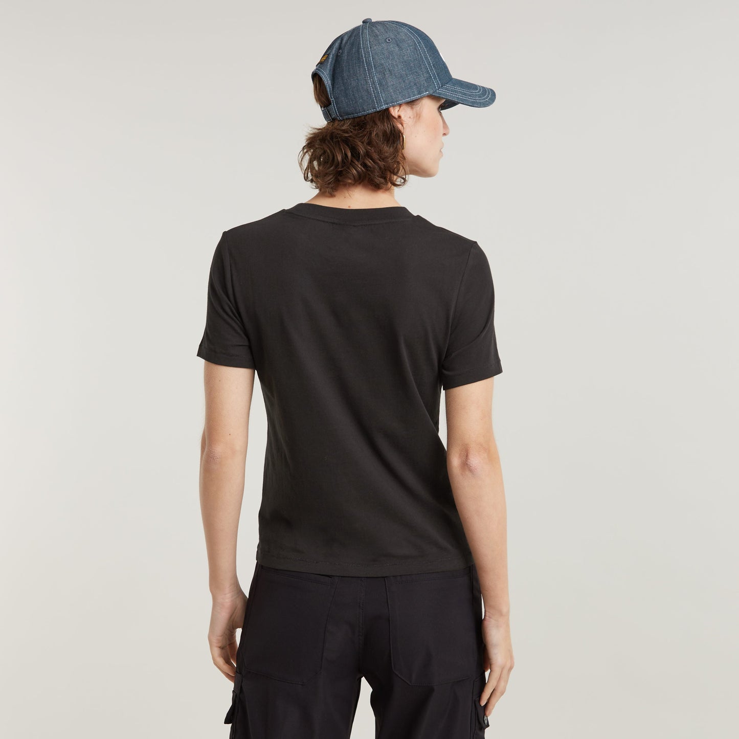 Modelo de espaldas luciendo el top con faux embroidered logo en color negro, complementado con una gorra de mezclilla.