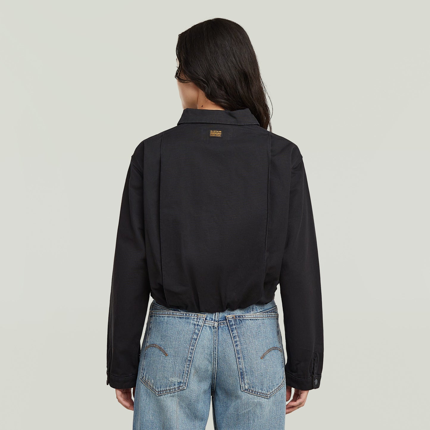 Back view of a woman wearing a black cropped barn jacket with denim jeans.