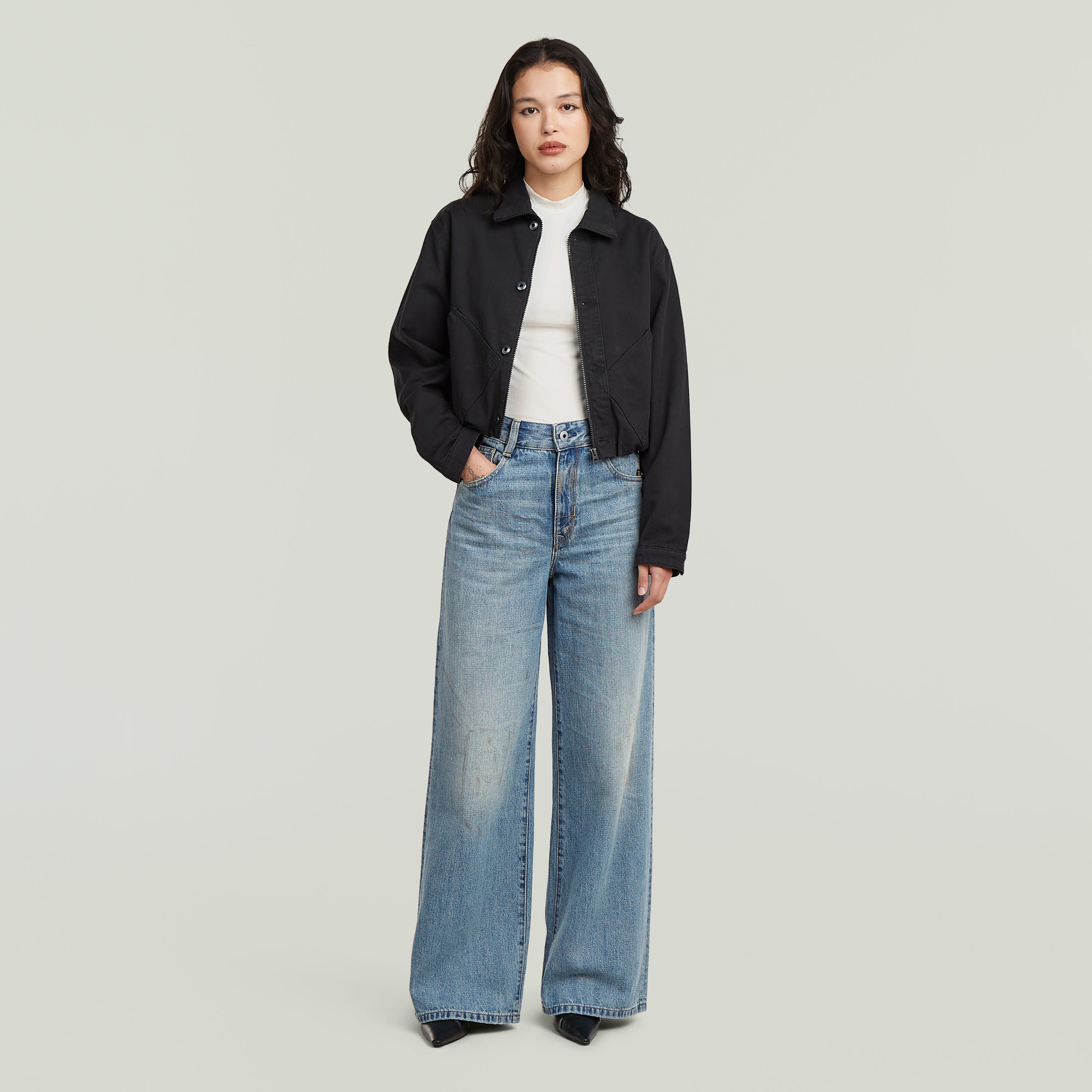 Mujer con chaqueta cropped barn negra y jeans anchos, posando en un fondo claro.
