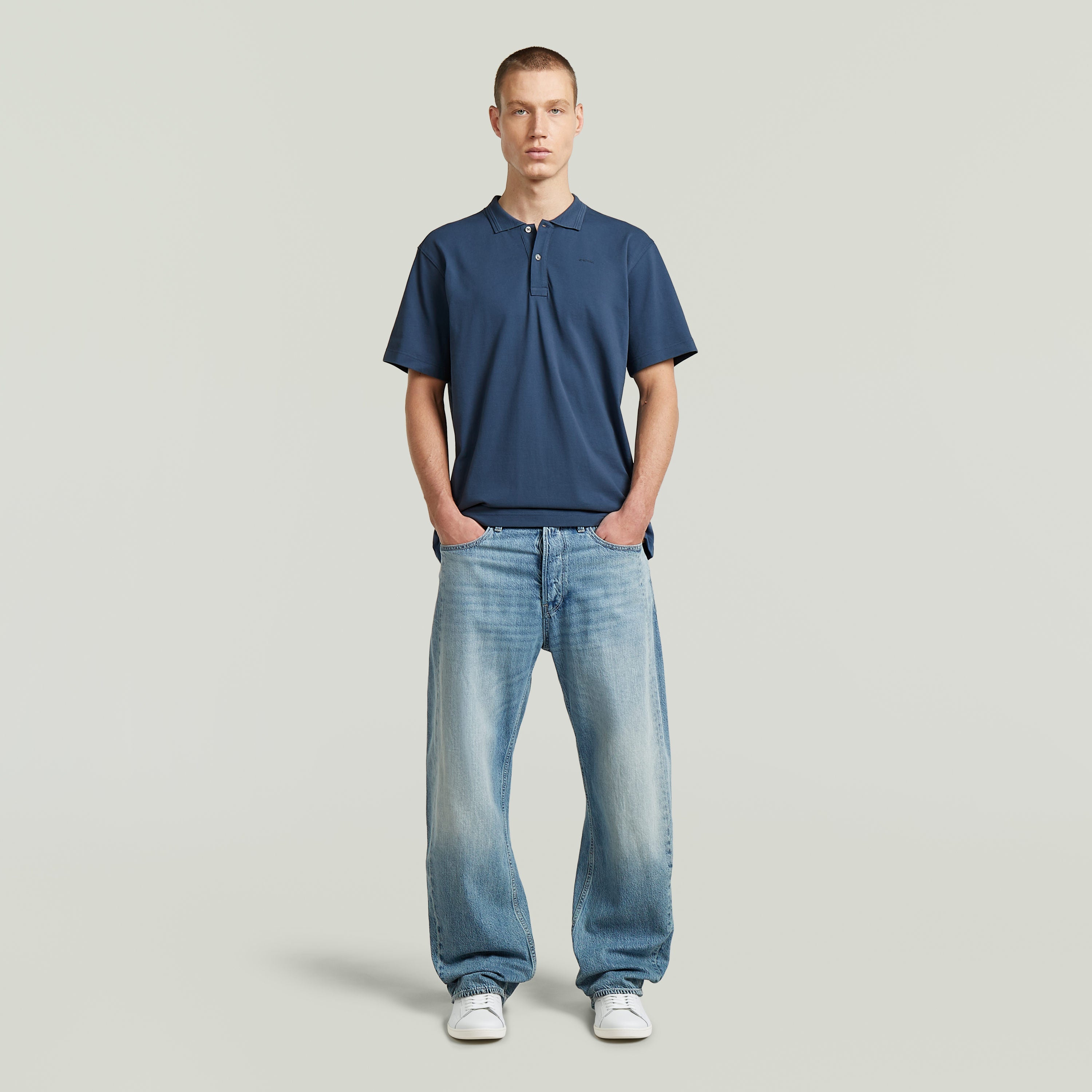 Hombre con camiseta polo core regular shirt de color azul intermedio y jeans, posando con las manos en los bolsillos.