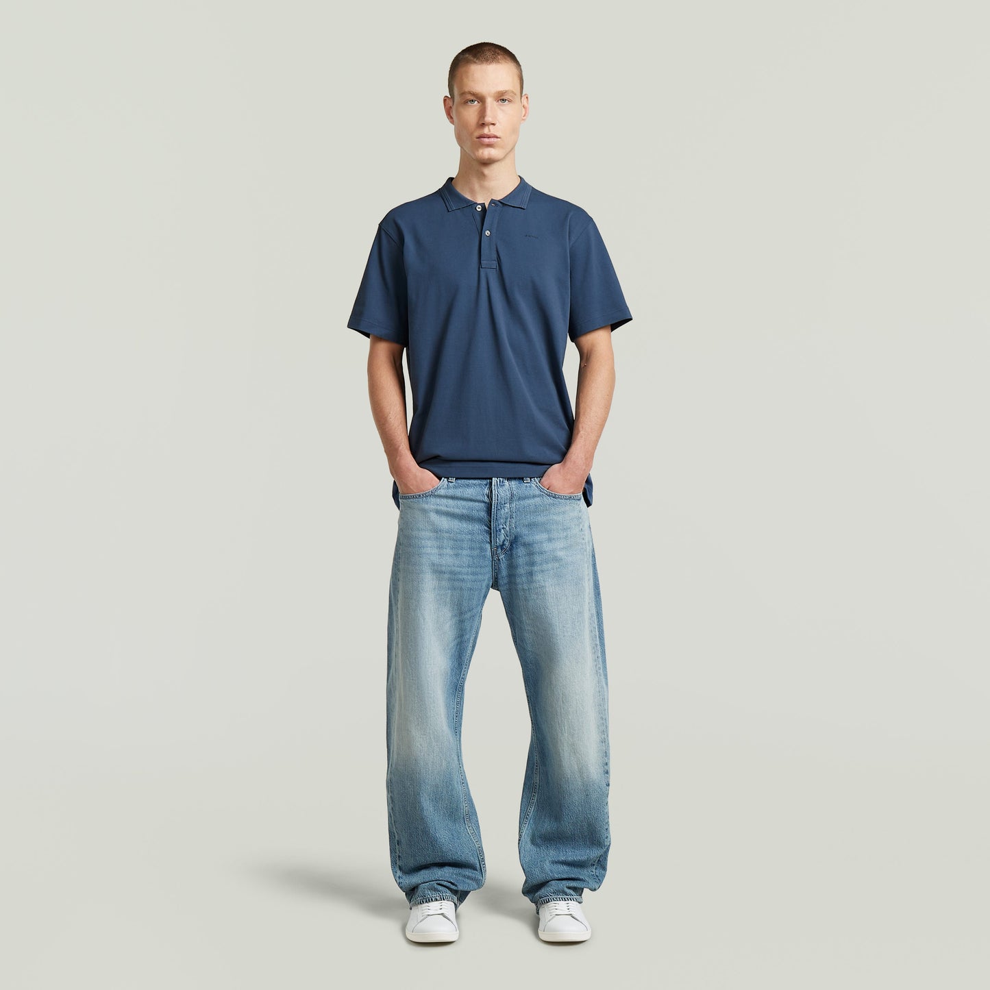 Hombre con camiseta polo core regular shirt de color azul intermedio y jeans, posando con las manos en los bolsillos.
