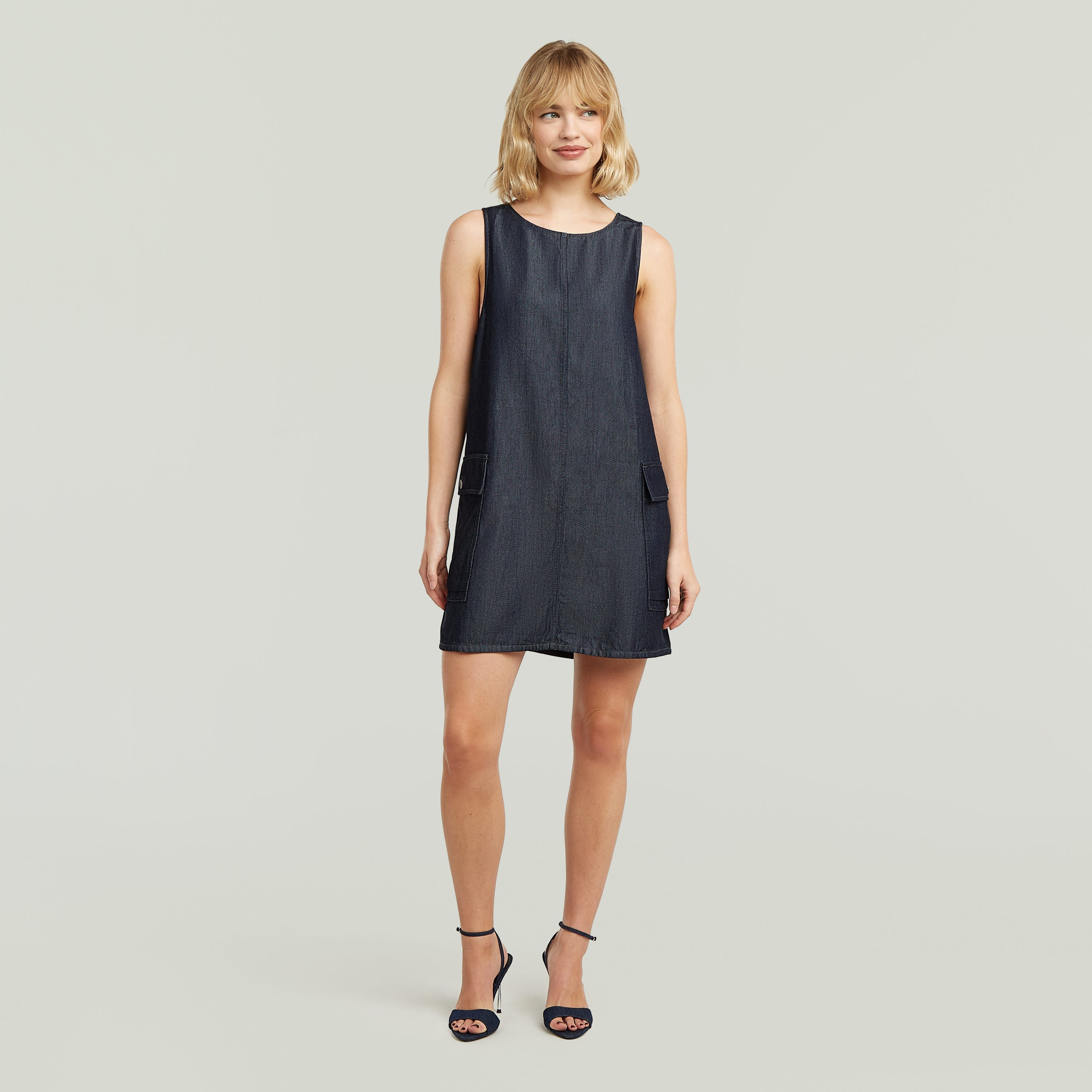 Modelo usando un vestido cargo shift ligero de denim oscuro con tirantes anchos y bolsillos laterales.