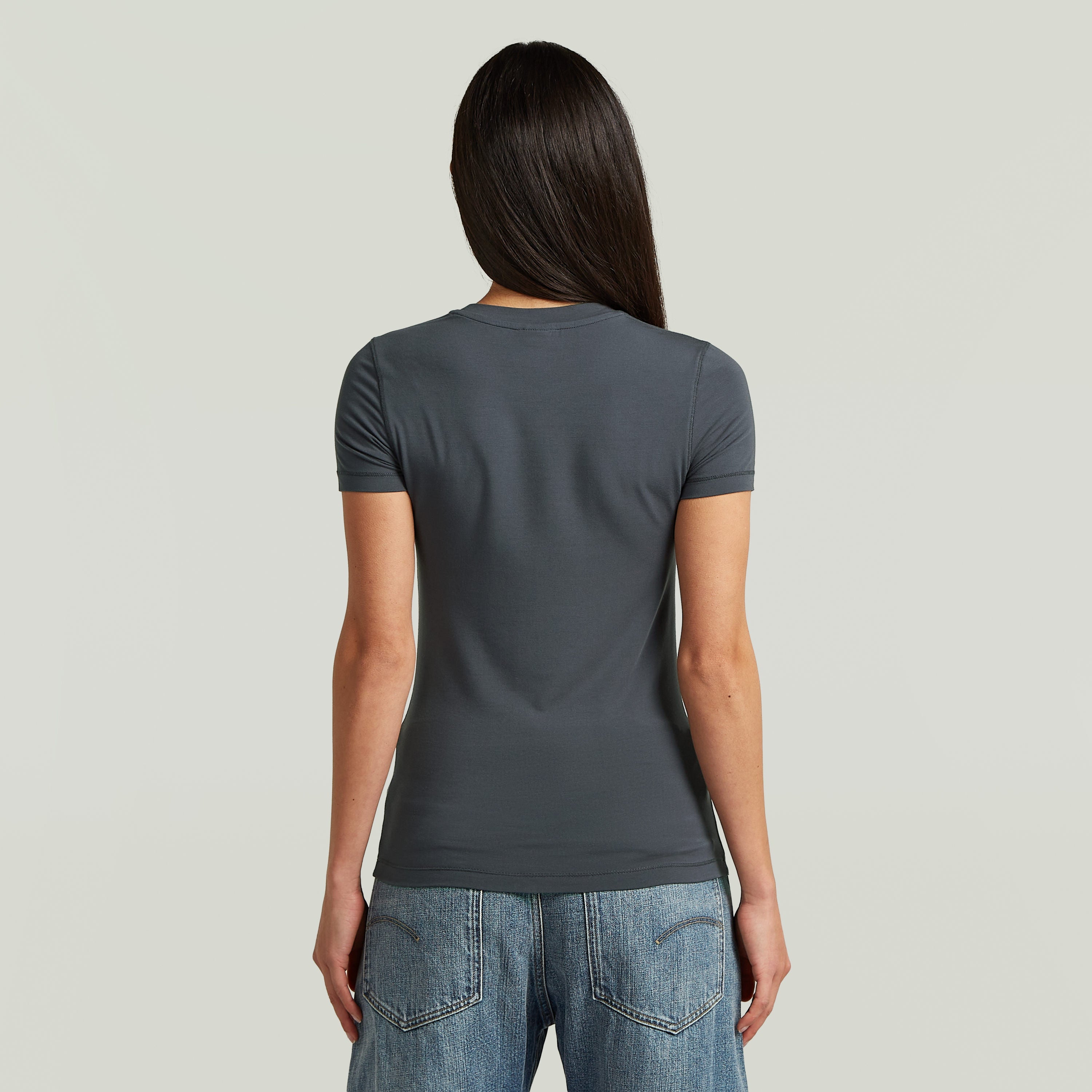 Mujer con camiseta 2nd Skin de corte ajustado, vista trasera, en color gris, combinada con jeans.