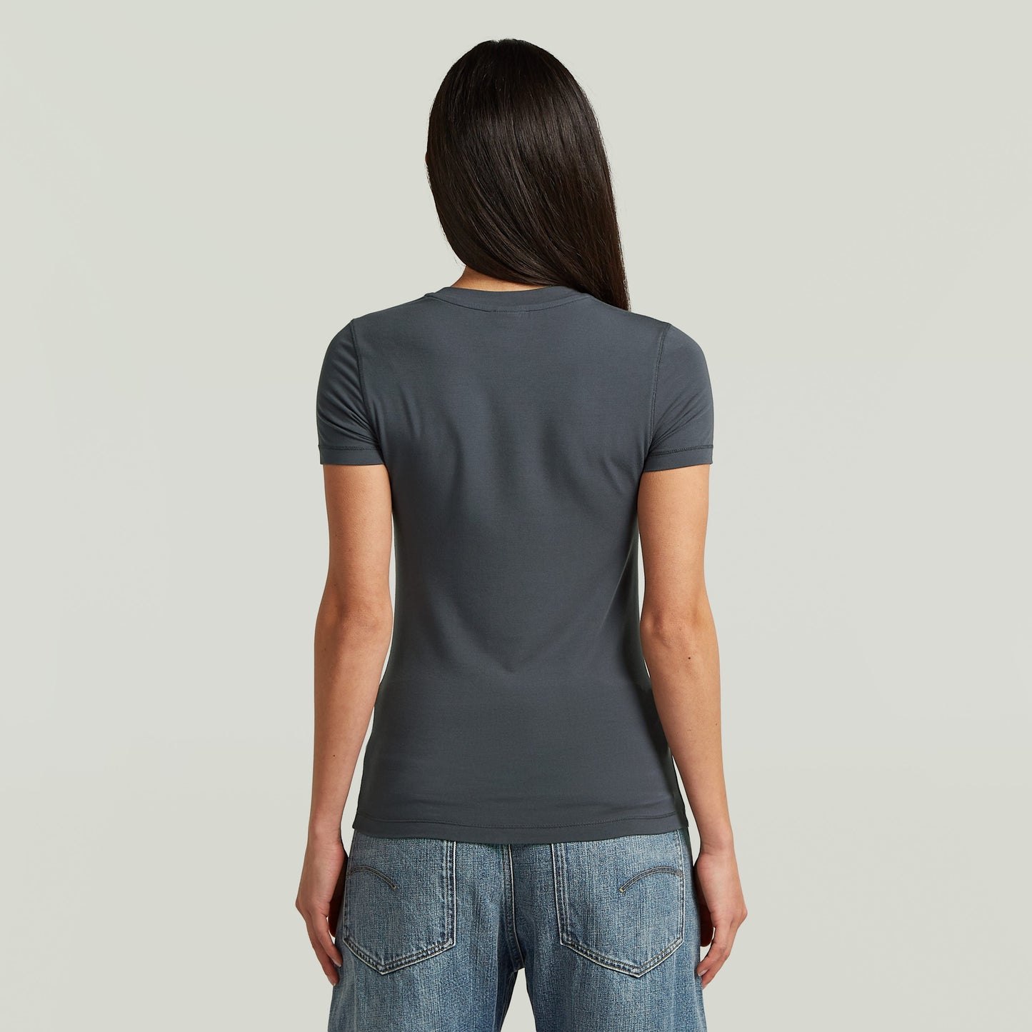Mujer con camiseta 2nd Skin de corte ajustado, vista trasera, en color gris, combinada con jeans.