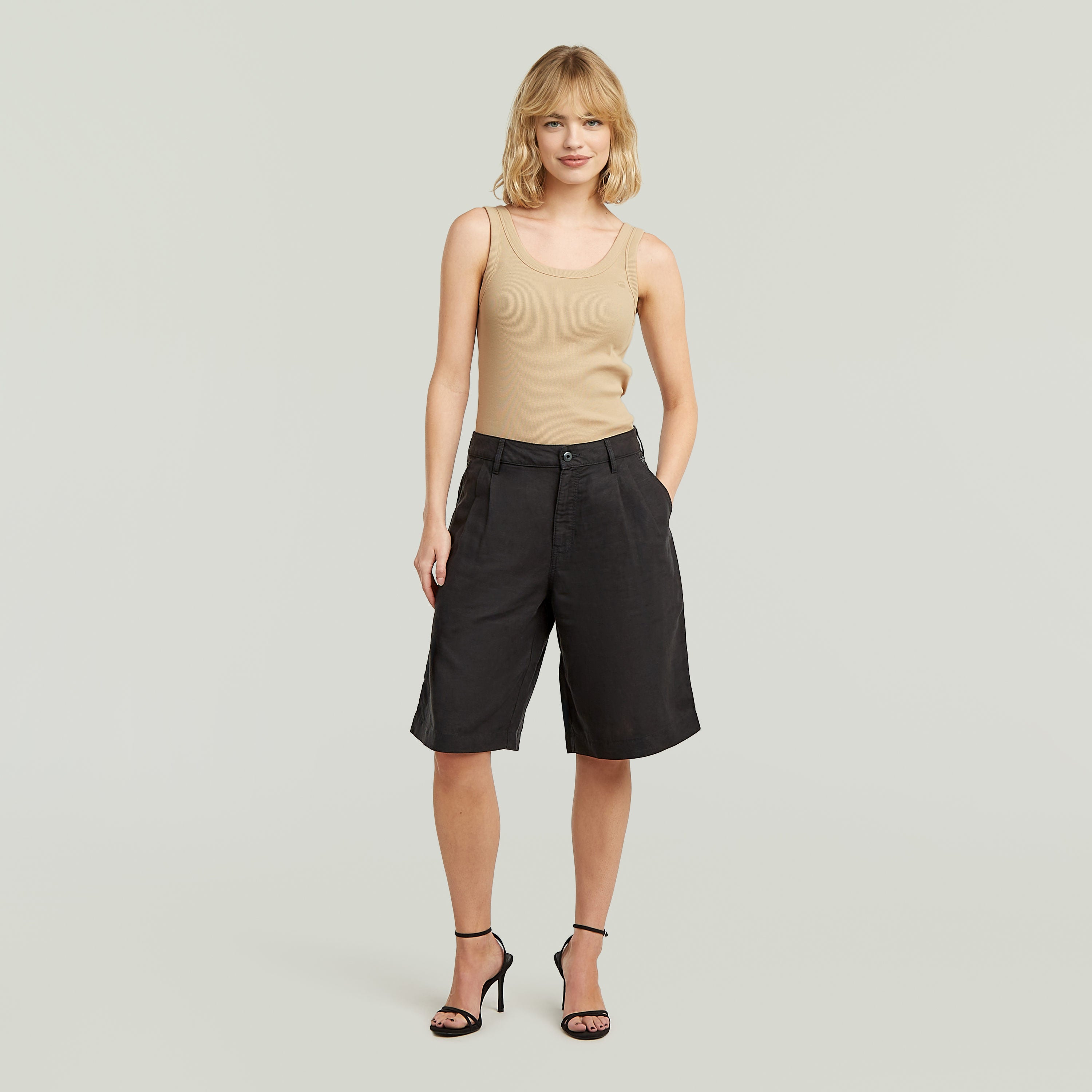 Mujer con shorts pleated bermuda negros, top beige, y tacones, fondo claro.
