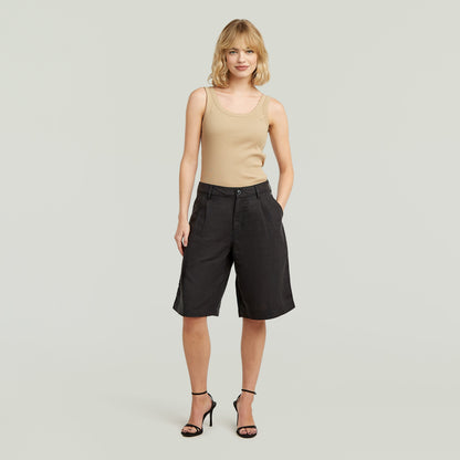 Mujer con shorts pleated bermuda negros, top beige, y tacones, fondo claro.