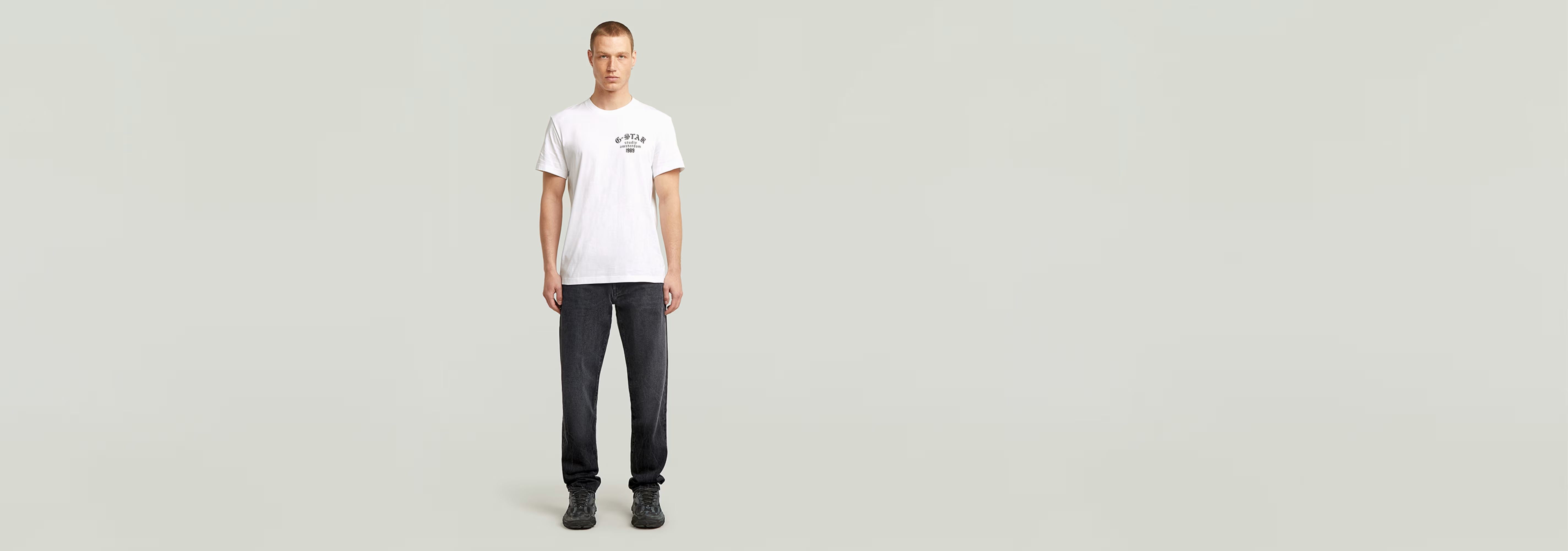 Hombre usando una camiseta Studio AMS blanca con diseño gráfico y pantalones oscuros sobre fondo claro.