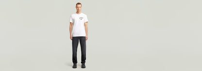 Hombre usando una camiseta Studio AMS blanca con diseño gráfico y pantalones oscuros sobre fondo claro.