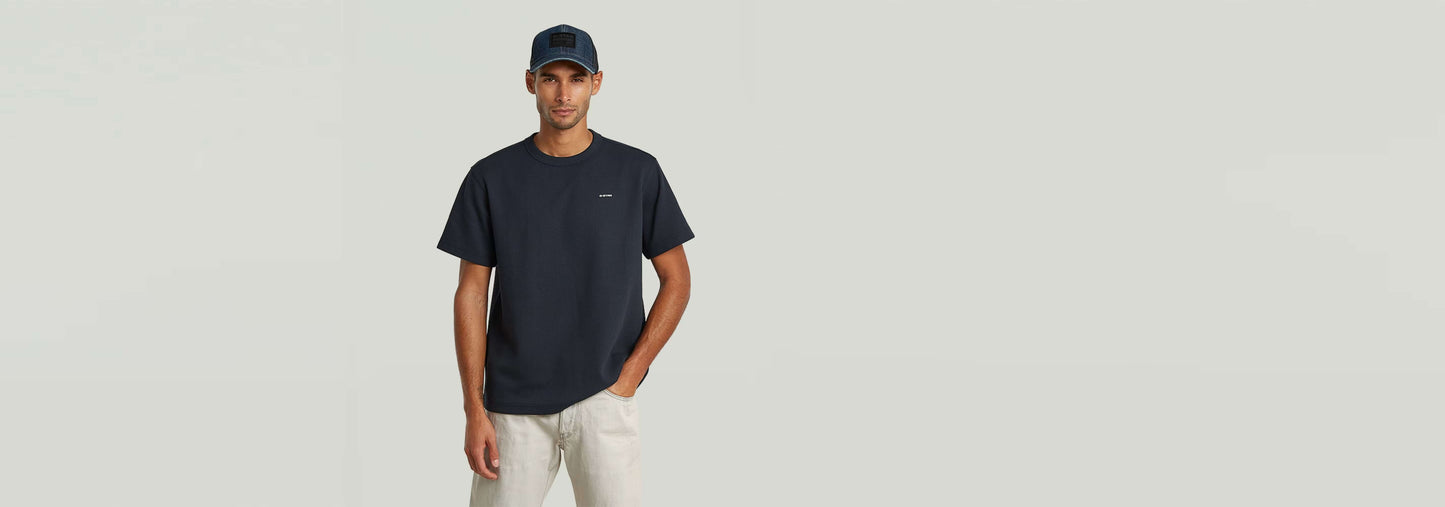 Hombre modelando una gorra de rejilla Baseball con camiseta oscura y pantalones claros en fondo neutro.