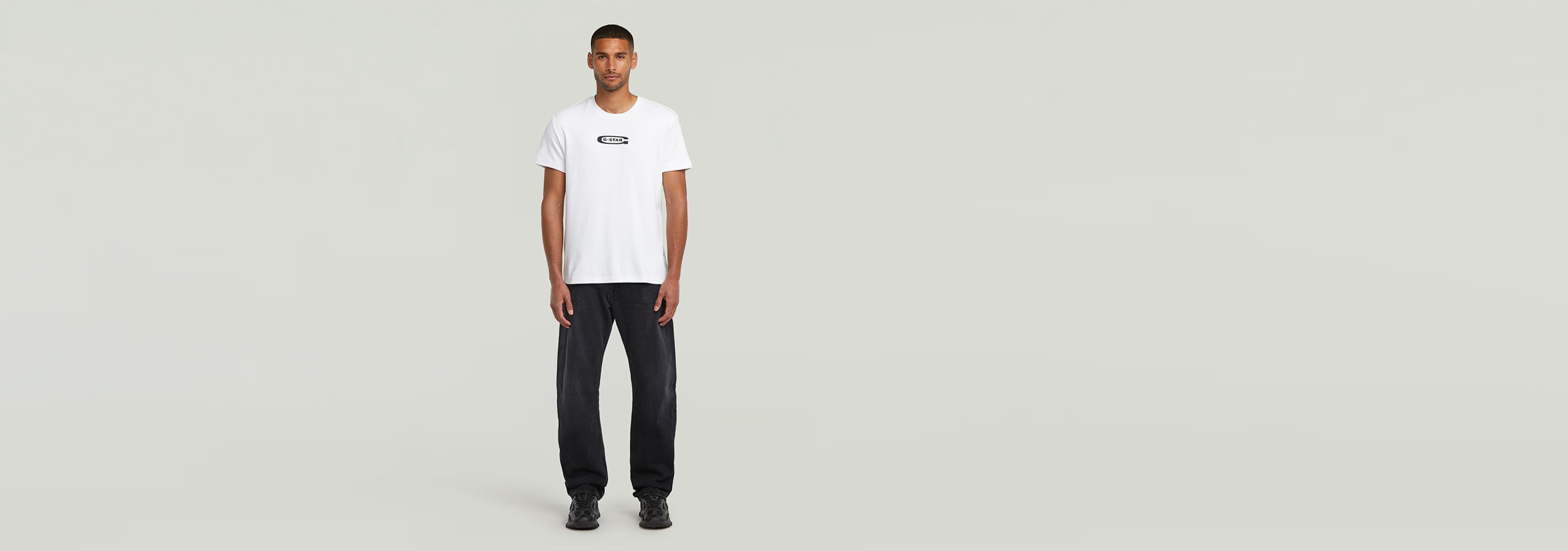 Modelo masculino vistiendo una camiseta blanca y pantalones oscuros, con logo en la parte frontal.