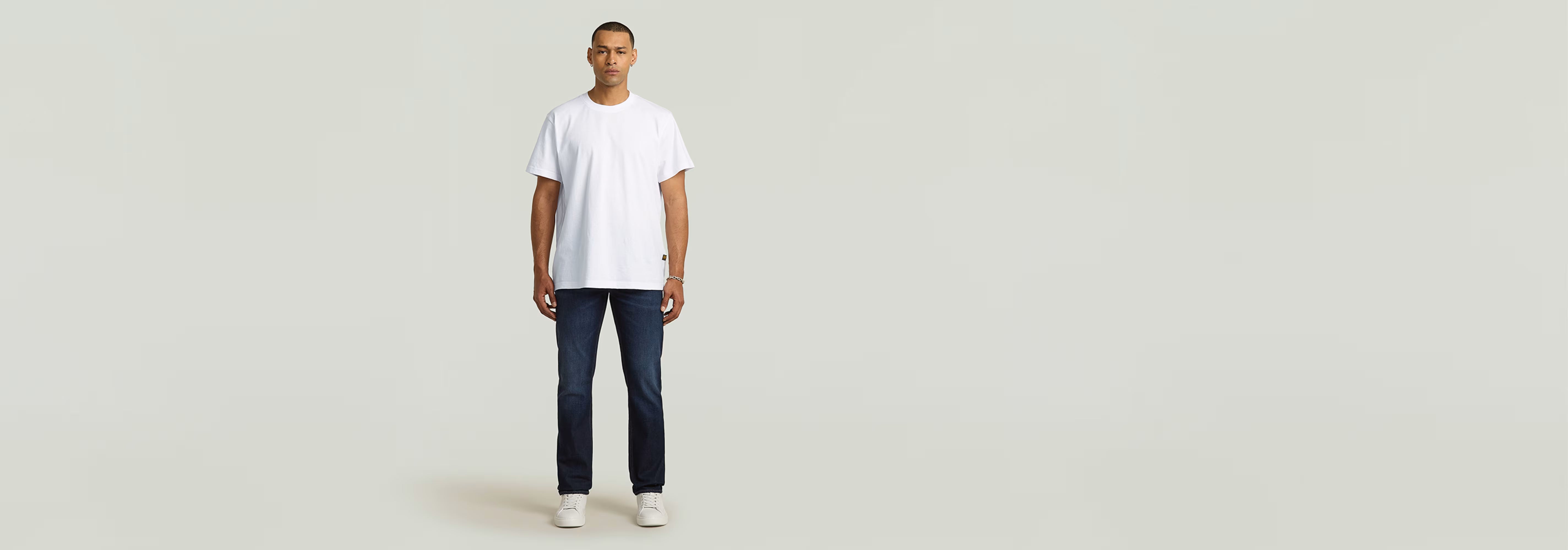 Modelo vistiendo Jeans Mosa Straight en un fondo neutro con camiseta blanca y zapatillas. Moda masculina.
