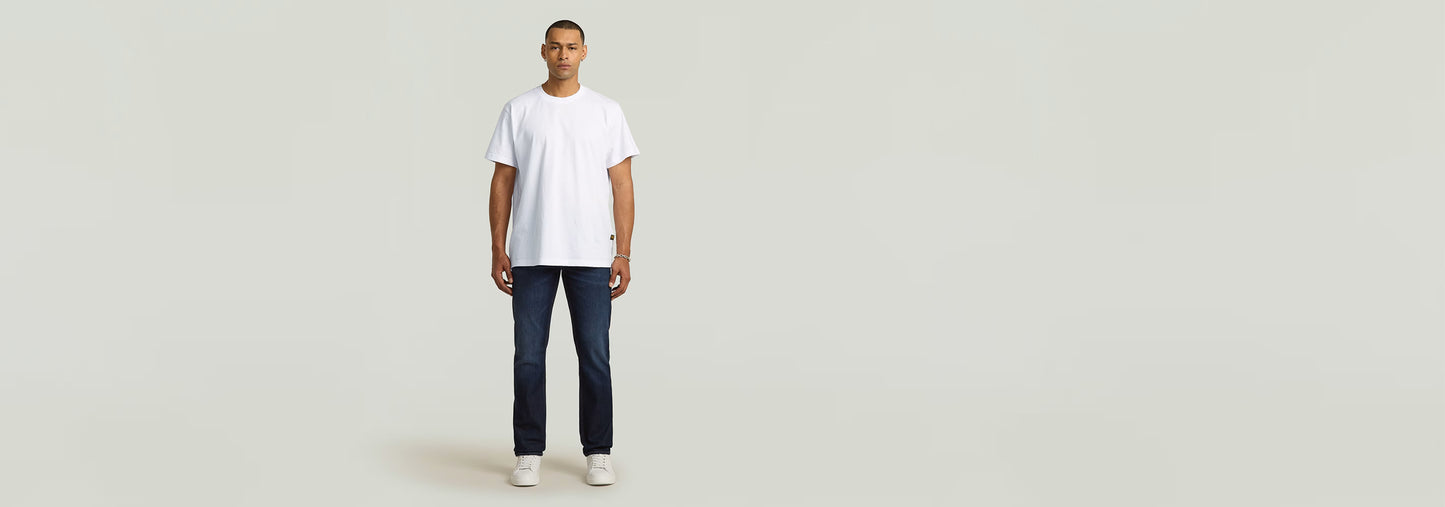 Modelo vistiendo Jeans Mosa Straight en un fondo neutro con camiseta blanca y zapatillas. Moda masculina.