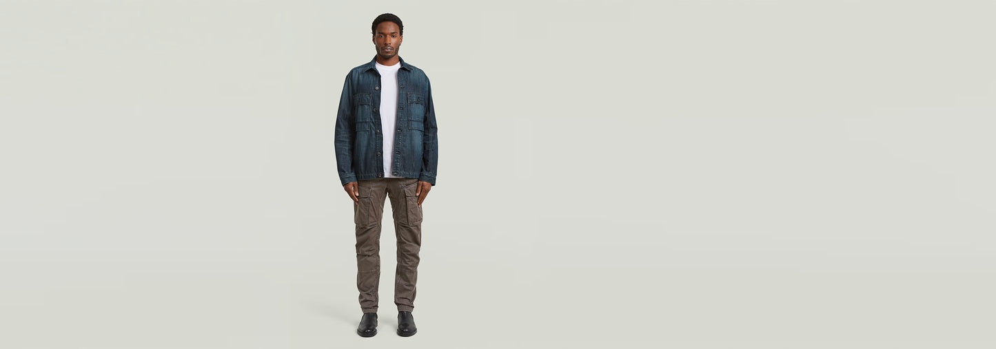 Modelo con Pantalones Rovic Zip 3D Regular Tapered, camiseta blanca y chaqueta azul, de pie sobre fondo claro.