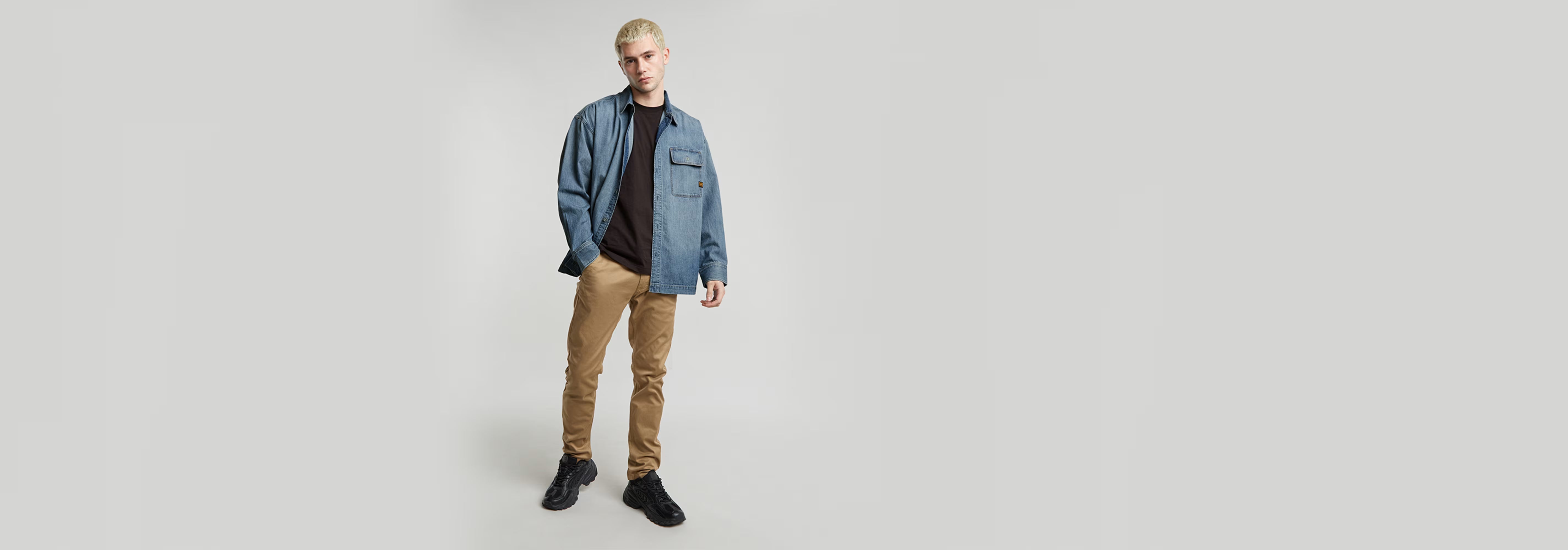 Pantalones Skinny Chino 3.0 en un modelo masculino con camisa y zapatos negros sobre fondo gris.