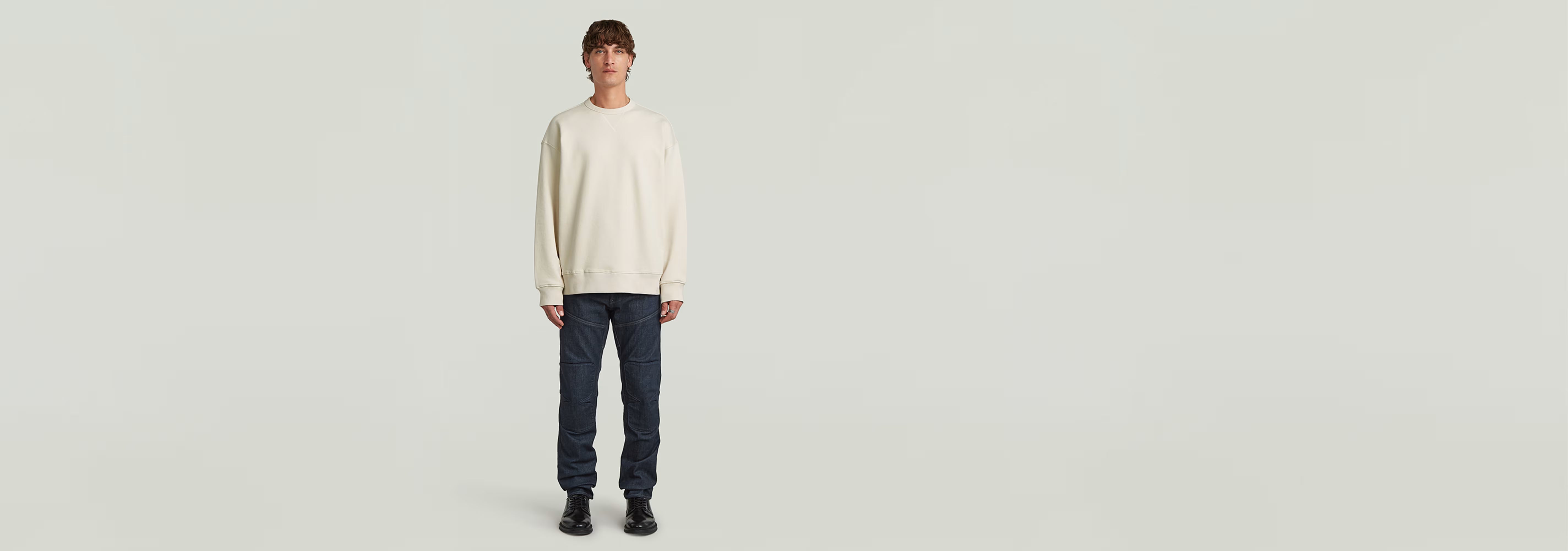 Hombre usando la Sudadera GS-01 Relaxed de color crema, combinada con jeans y zapatos negros en un fondo claro.