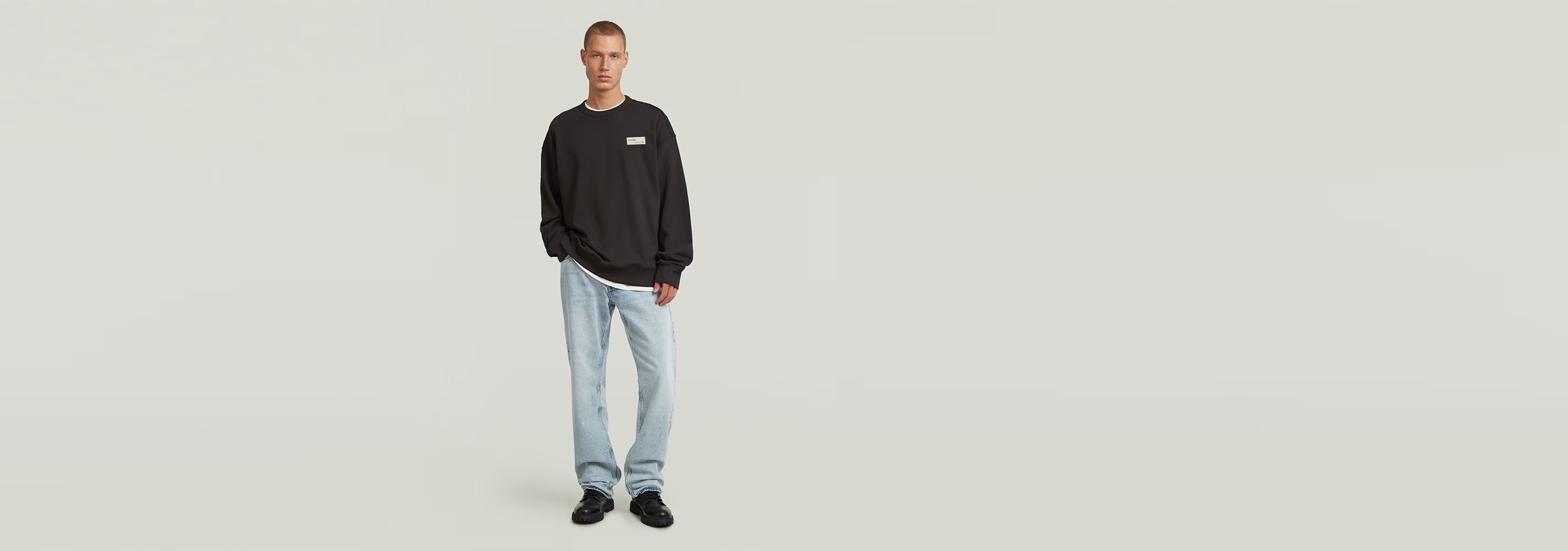 Hombre usando sudadera HB Badge de corte relajado, con jeans claros y zapatillas, fondo blanco.